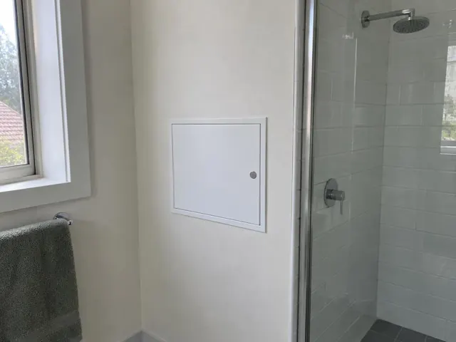 Bathroom wall with a small access panel beside a glass shower and window in Stanford Merthyr